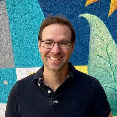Smiling man wearing glasses and a black polo shirt standing in front of a colorful mural with blue, yellow, and green shapes.
